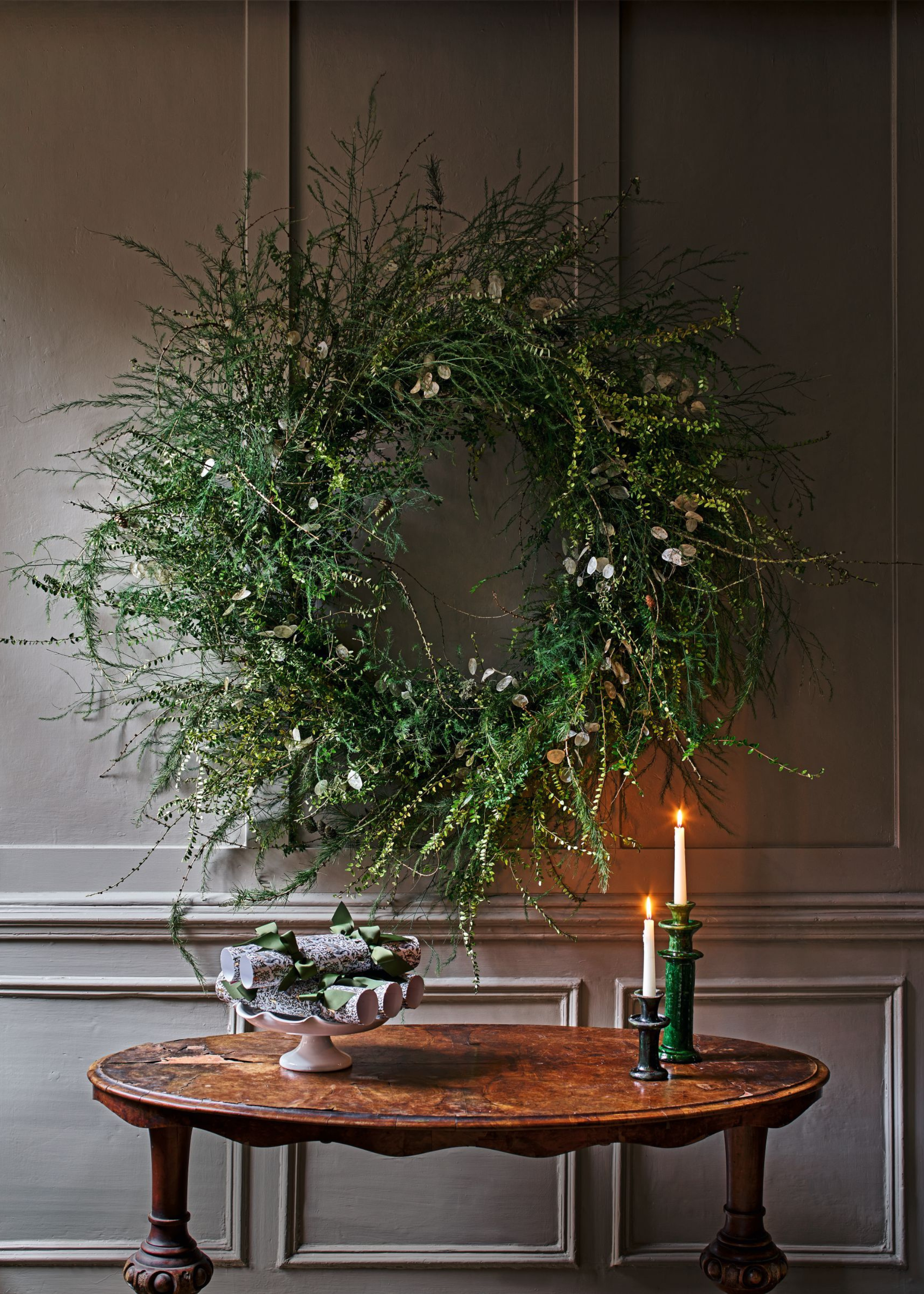 An oval hallway table has two taper candles lit next to a bowl filled with christmas crackers with a large wild wreath on the wall behind