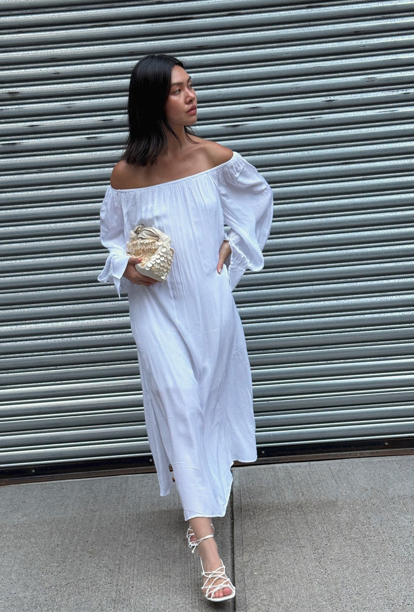 The coastal boho trend is shown in a photo of a woman standing on the sidewalk wearing a white off-the-shoulder maxi dress styled with white strappy sandals, and a seashell clutch bag