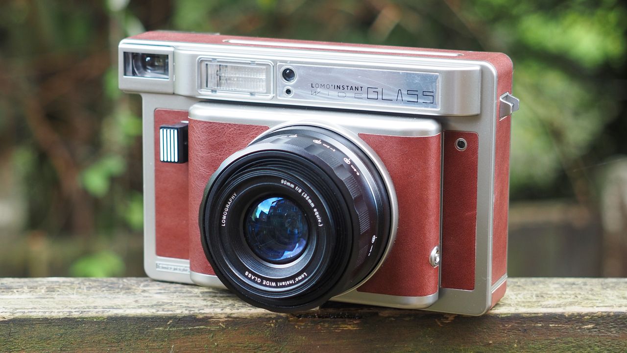 Lomo'Instant Wide Glass camera, sitting on a wooden surface, outdoors against a woodland background
