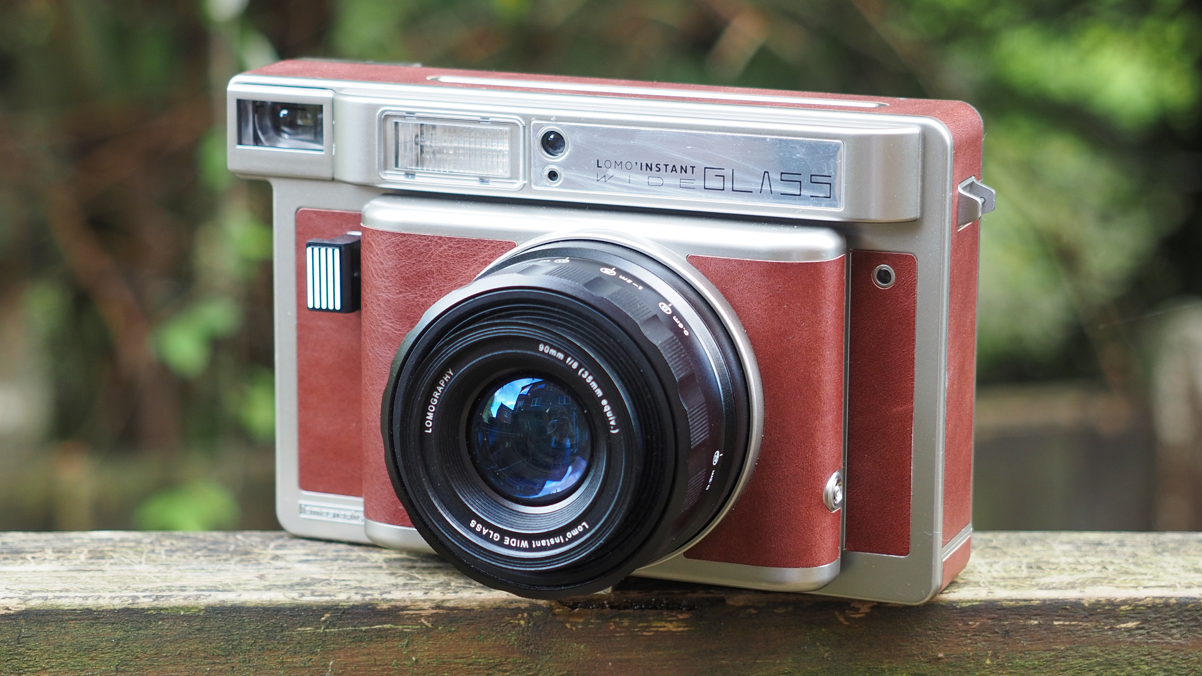 Lomo'Instant Wide Glass camera, sitting on a wooden surface, outdoors against a wooded background