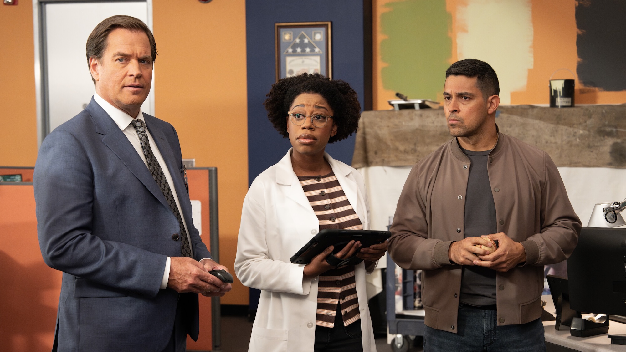 Tony Dinozzo in blue-gray suit standing with Kasie Hines in labcoat and striped shirt, holding a tablet, and Nick Torres in brown jacket, all standing in NCIS office in NCIS Season 24