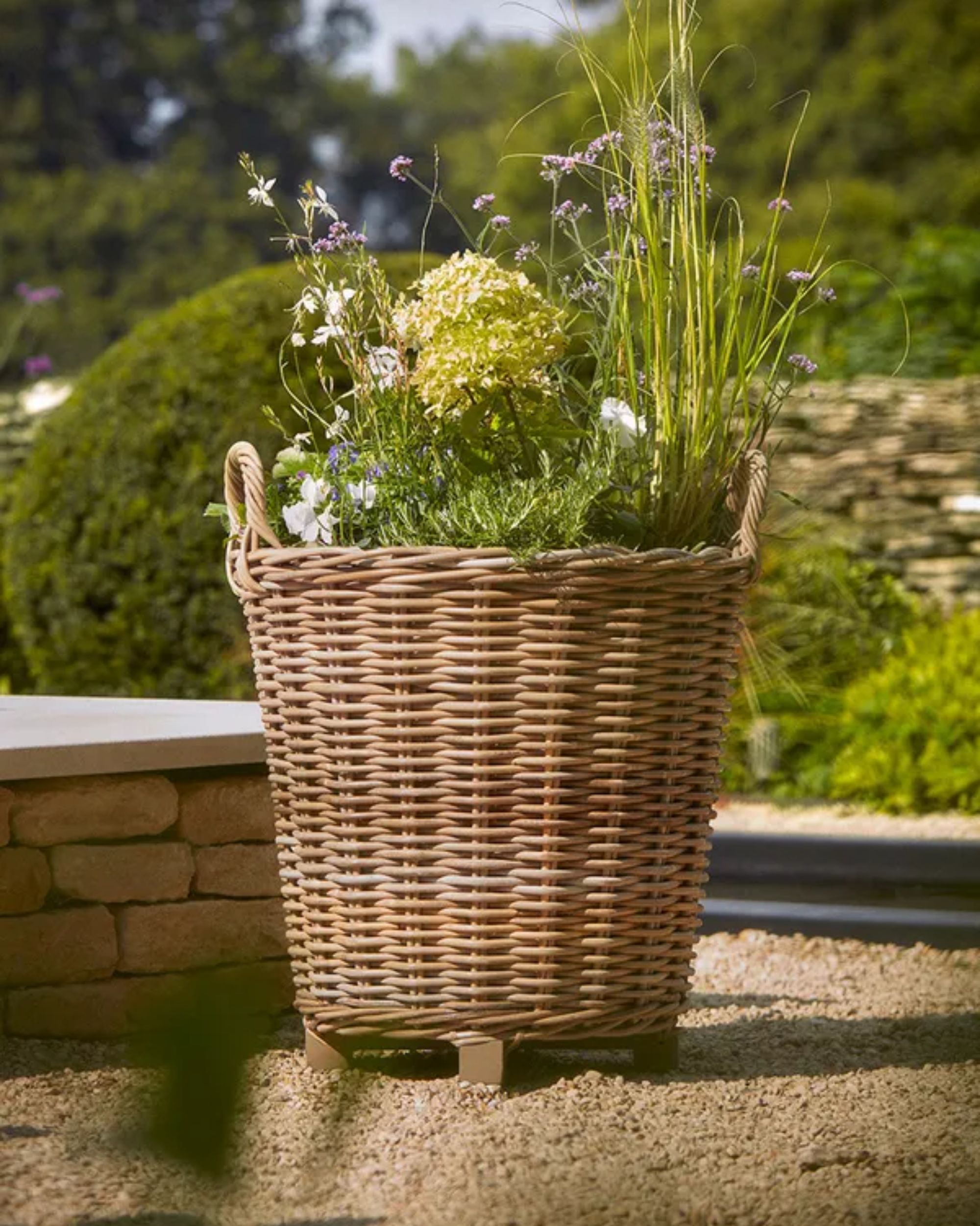 A rattan planter outside with green and lilac flowers planted inside.