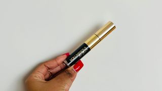 A person's hand holding a black mascara tube with a gold top, for L'oreal Voluminous Noir Balm Mascara review.