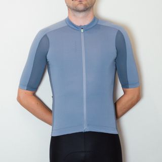 A white man in a pale blue cycling jersey and black cycling shorts stands against a white wall