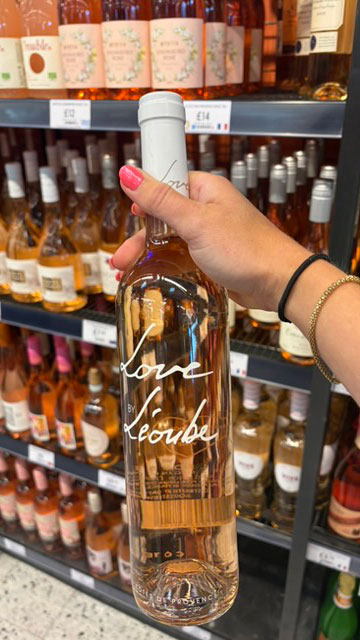 A hand holding a bottle of Love L&amp;eacute;oube Provence ros&amp;eacute; with shelves of ros&amp;eacute; in the background