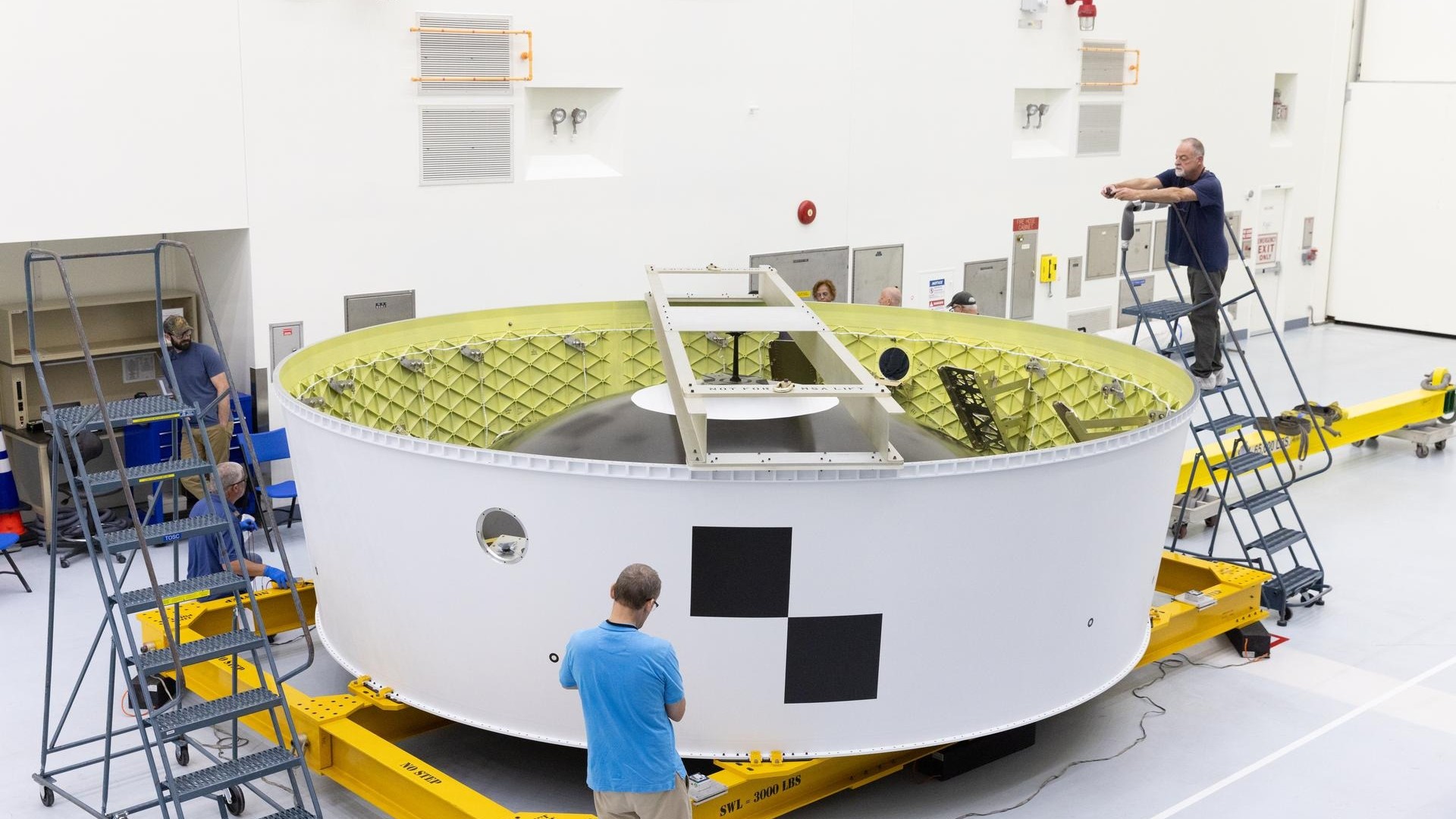 a large white metal ring sits on a large yellow metal dolly in a hangar surrounded by eight people