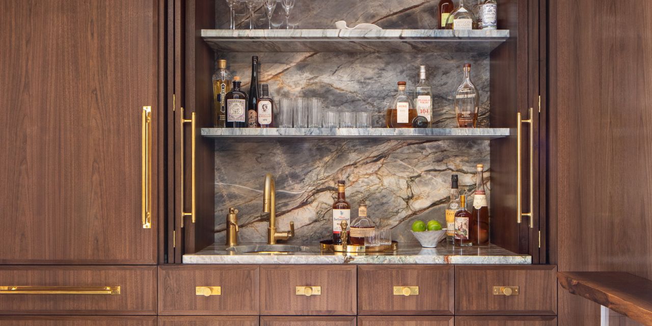 Close-up image of a home closet bar. The cabinetry has a walnut wood finish and the shelves and backsplash inside of the bar are gray, brown, and gold marble. There are gold hardware accents as well.