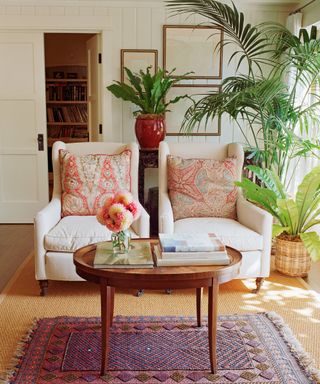 Two white upholstered armchairs with coral, patterned back cushions in grand living room with round wooden coffee table, patterned rug and lots of houseplants