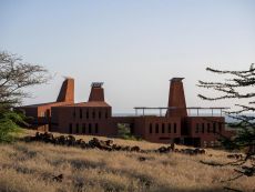 The education campus is surrounded by nature. There are three connected buildings, in burnt brown color. Each building has an element with an opening at the end, that looks like it's protruding from the building.