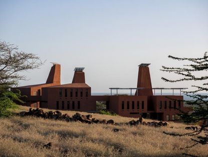 The education campus is surrounded by nature. There are three connected buildings, in burnt brown color. Each building has an element with an opening at the end, that looks like it's protruding from the building.