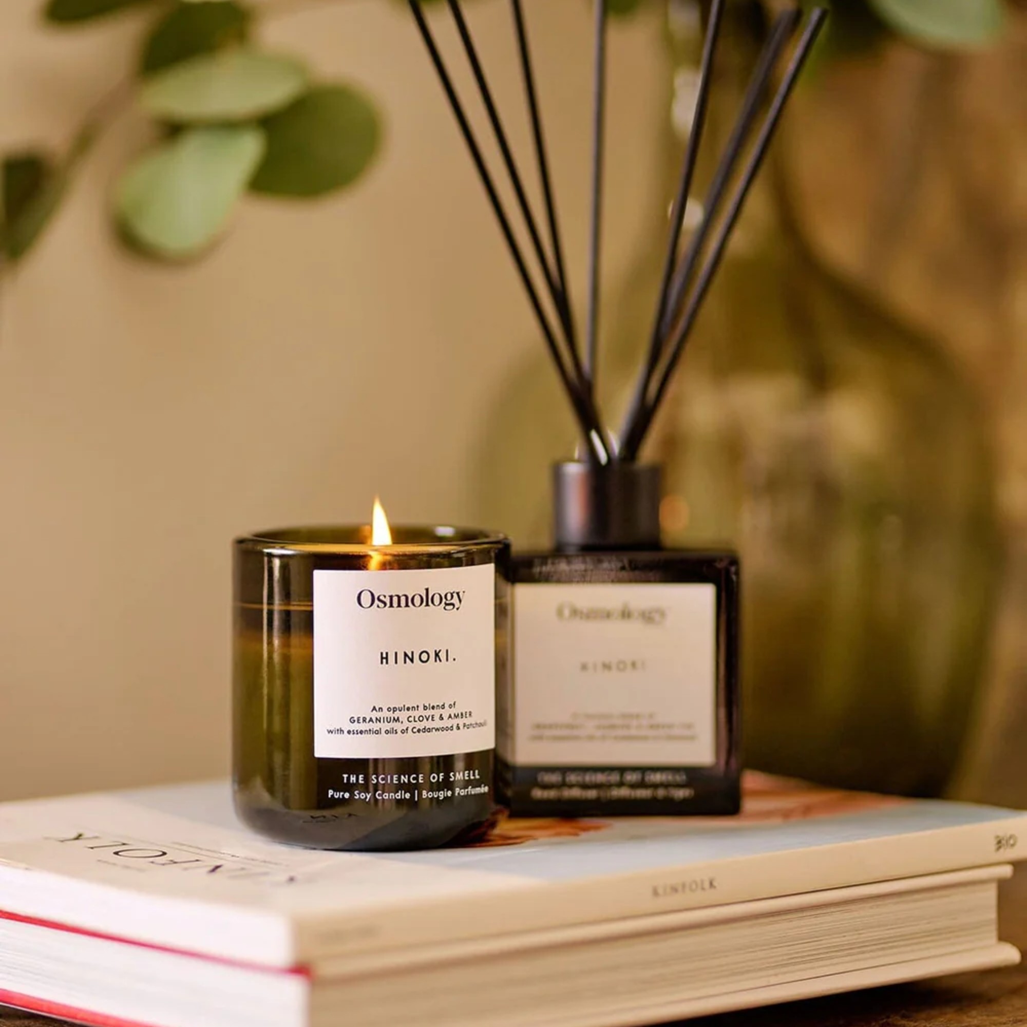 A lit Hinoki scented candle from Osmology with a matching reed diffuser on top of a book and a magazine