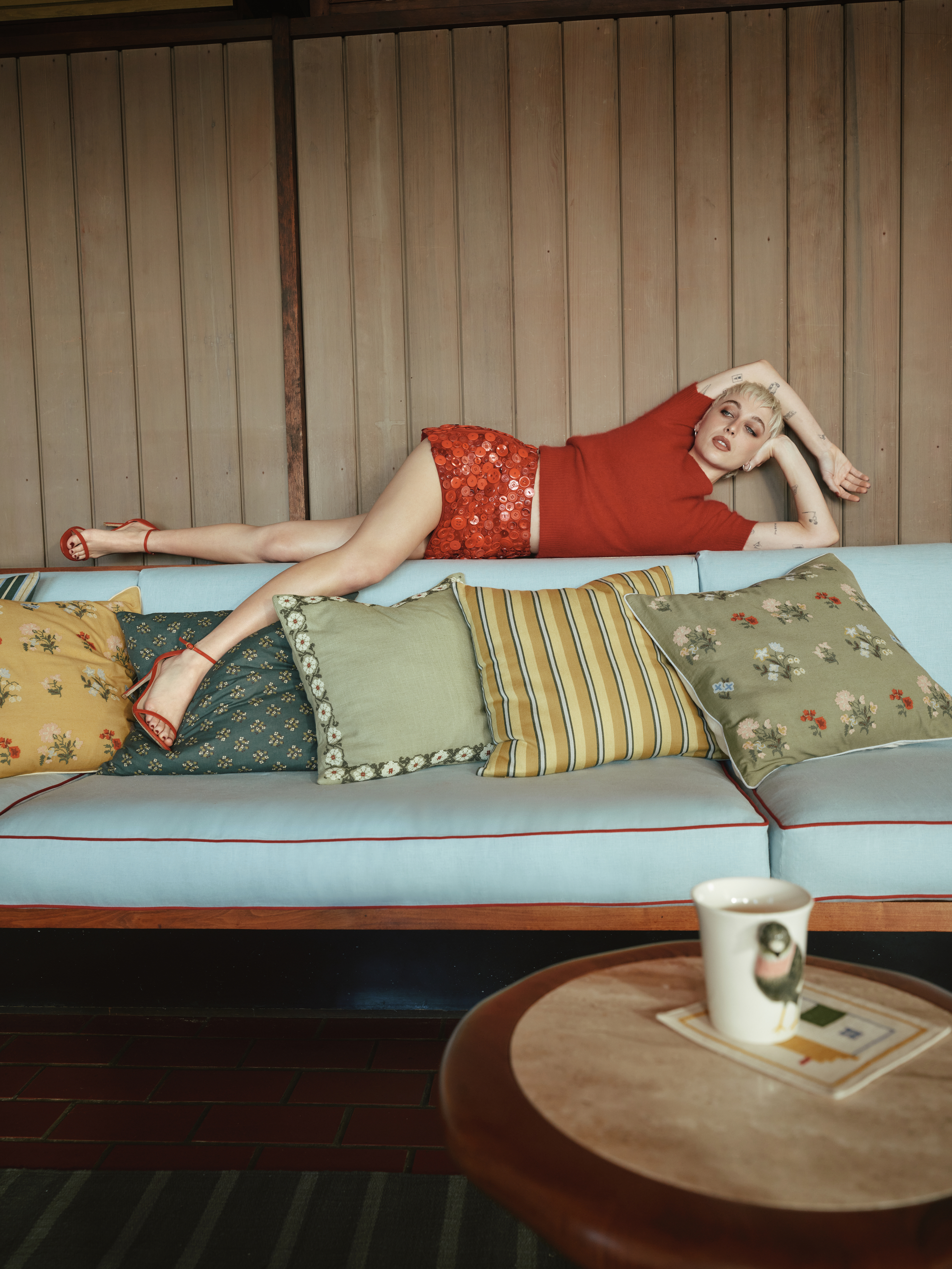 Image of a woman in a red outfit laying across the top of a light blue sofa with multiple green and yellow pillows on it.