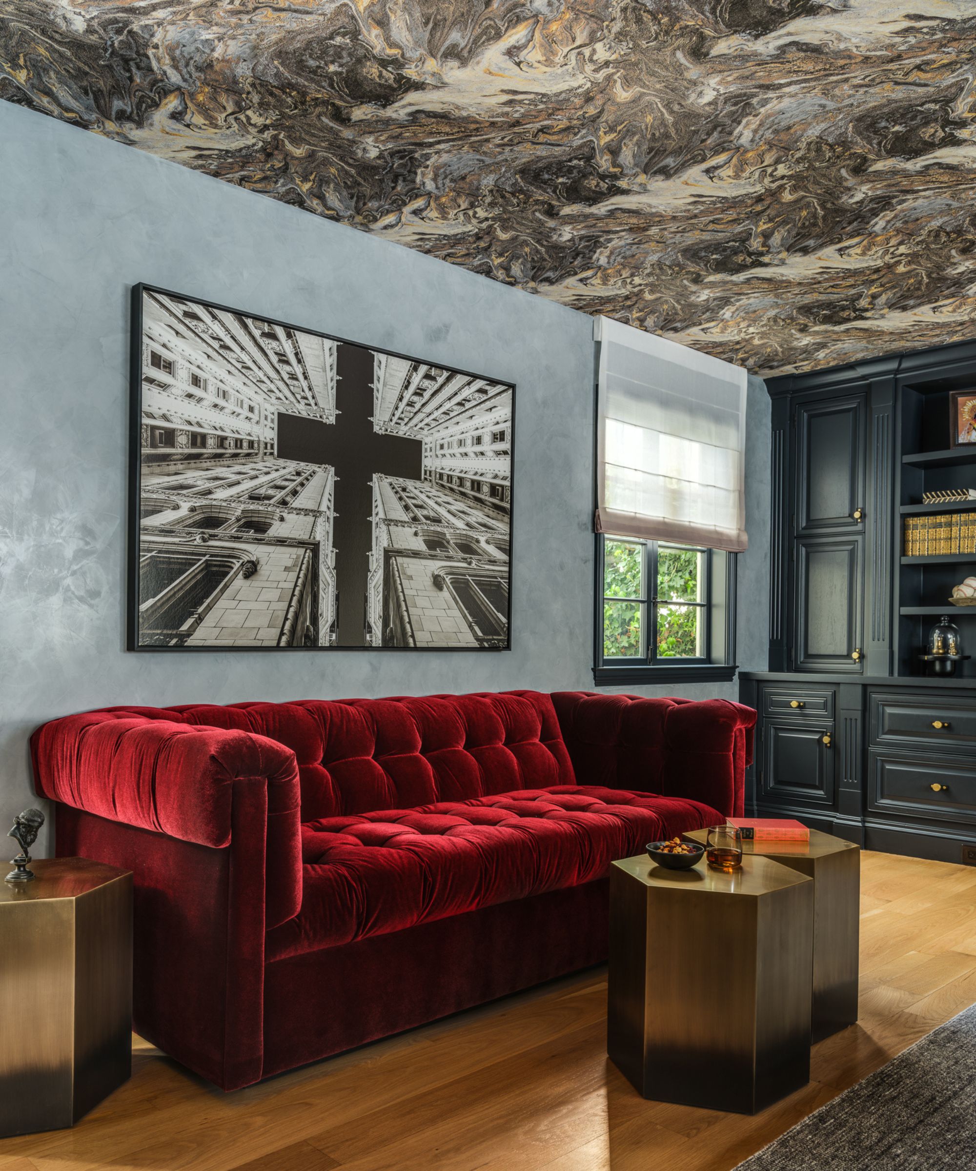 Bold living room with red velvet sofa, blue walls, marble wallpapered ceiling, three gold side tables. Blue painted cabinetry decorated with books. Large painting hung above sofa.