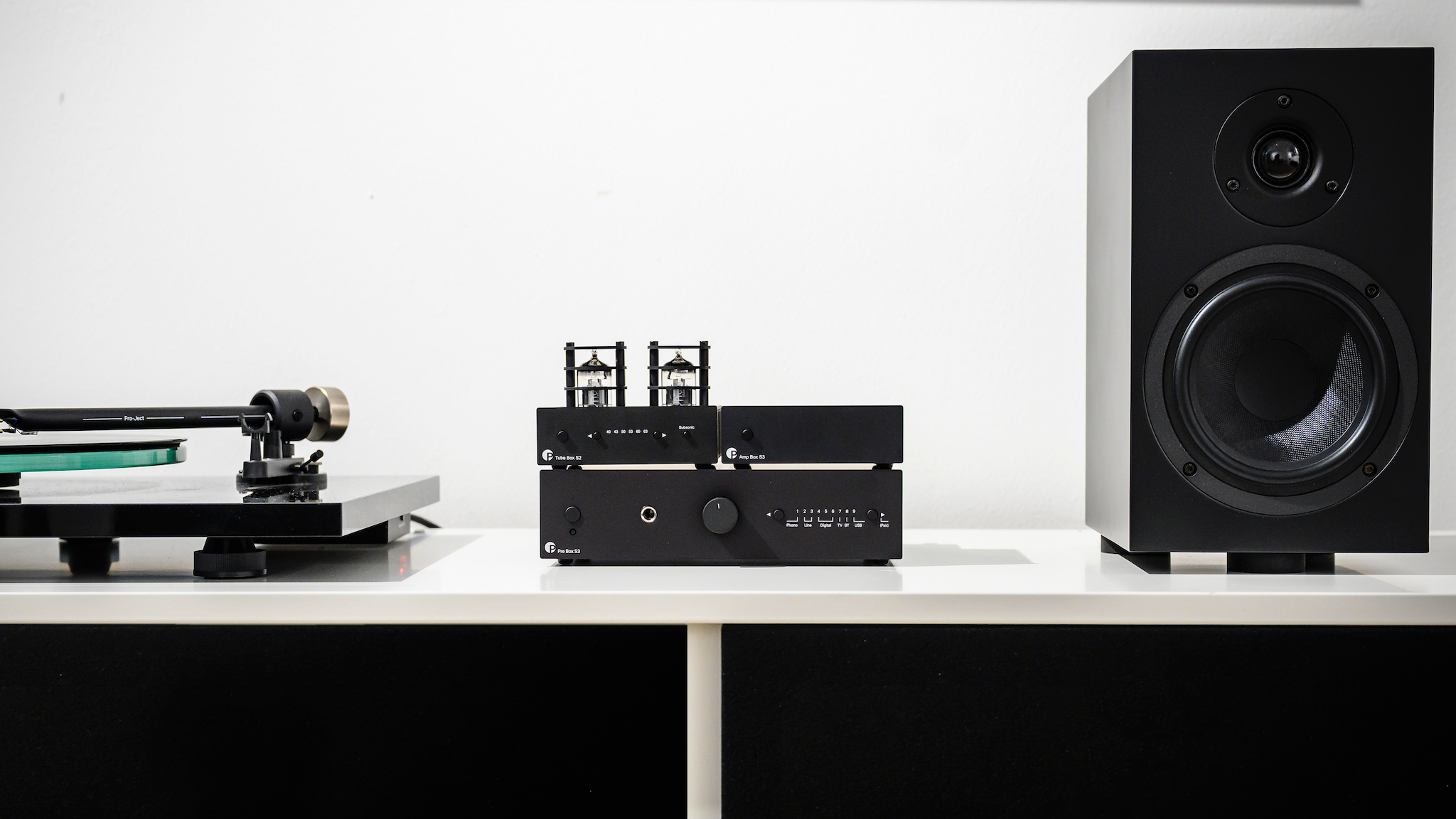 Pro-Ject Pre Box S3 underneath a Pro-Ject tube amplifier, between a Pro-Ject turntable and speaker against a white wall.