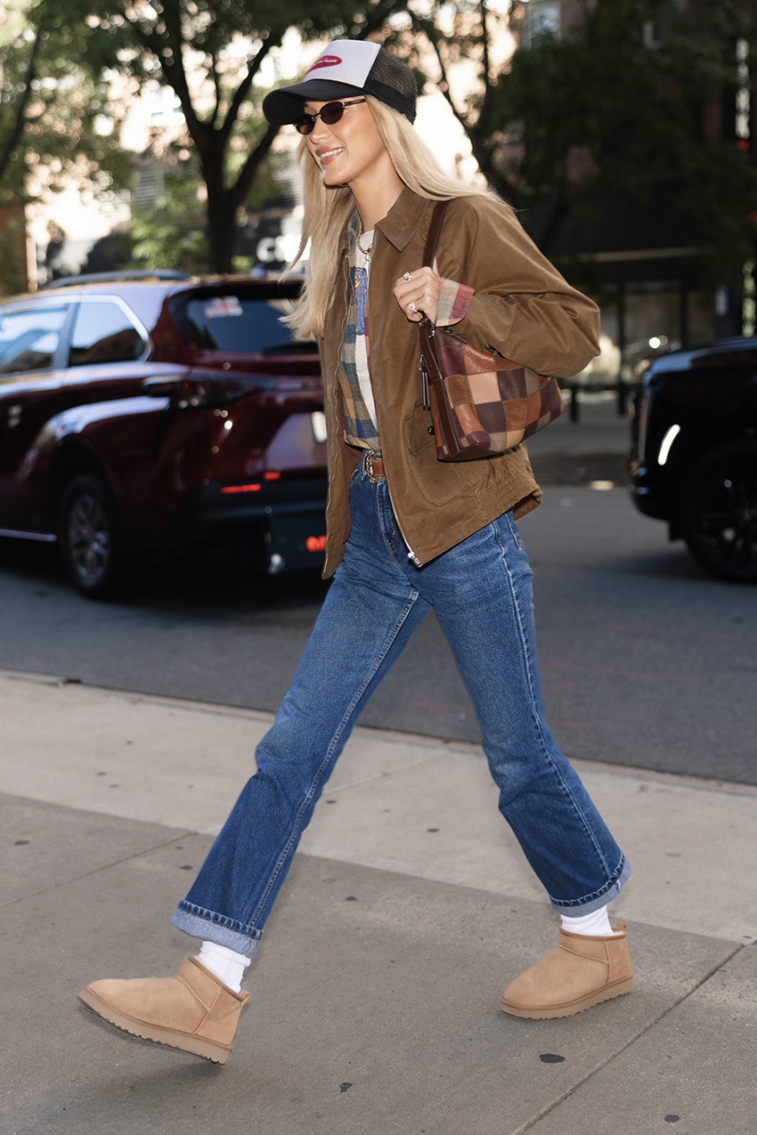 Bella Hadid wearing a brown collared jacket, plaid shirt over a T-shirt, and jeans with a baseball cap, Coach Alter/Ego Slouchy Shoulder Bag, and Ugg Classic Ultra Mini Boots with white socks.