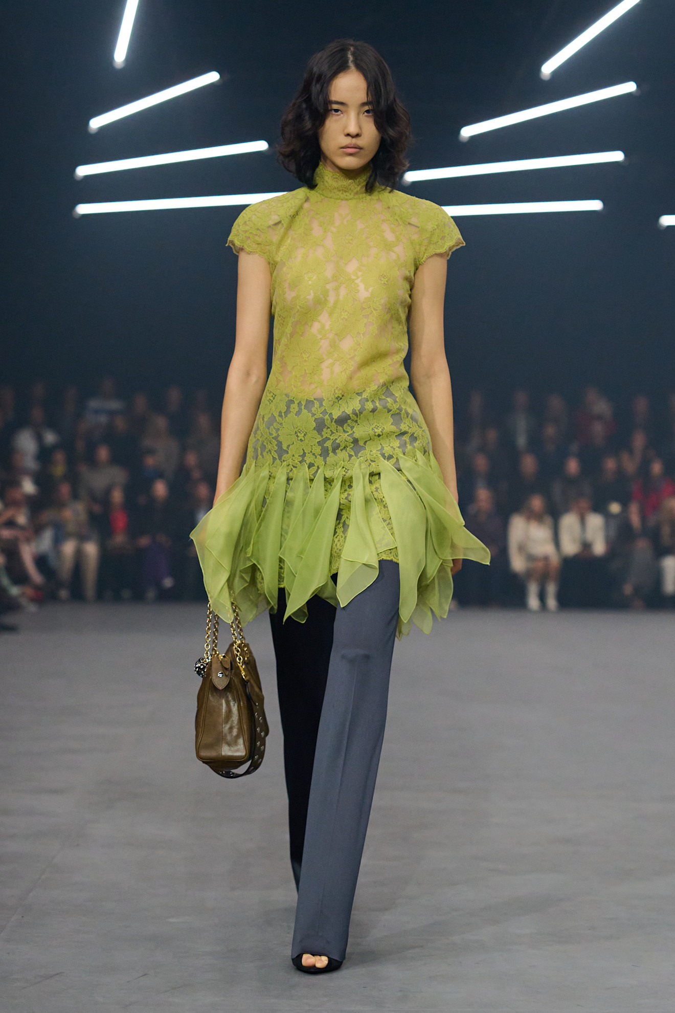 A Valentino model wearing a bright green sheer lace blouse with ruffles, gray slim trousers, black peep-toe sandals, and an olive green handbag with a two-tone chain strap.