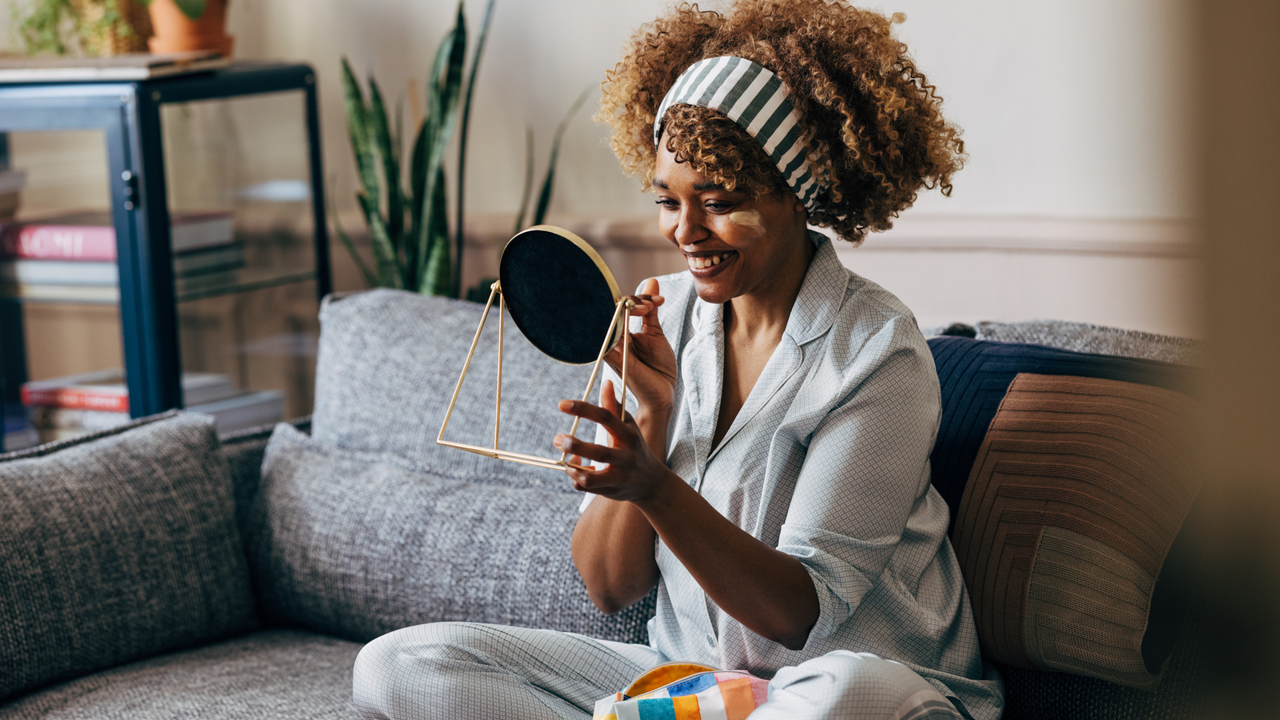 A wide angle view of a smiling African-American female holding a mirror while applying some creme on her face to keep it fresh and hydrated. She is sitting on the sofa in the living room