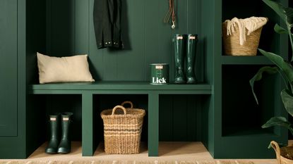 Image of a hunter green built-in mud room unit. There are two pairs of hunter green hunter boots, woven baskets, a can of Lick paint, a beige pillow, and a jacket hanging.