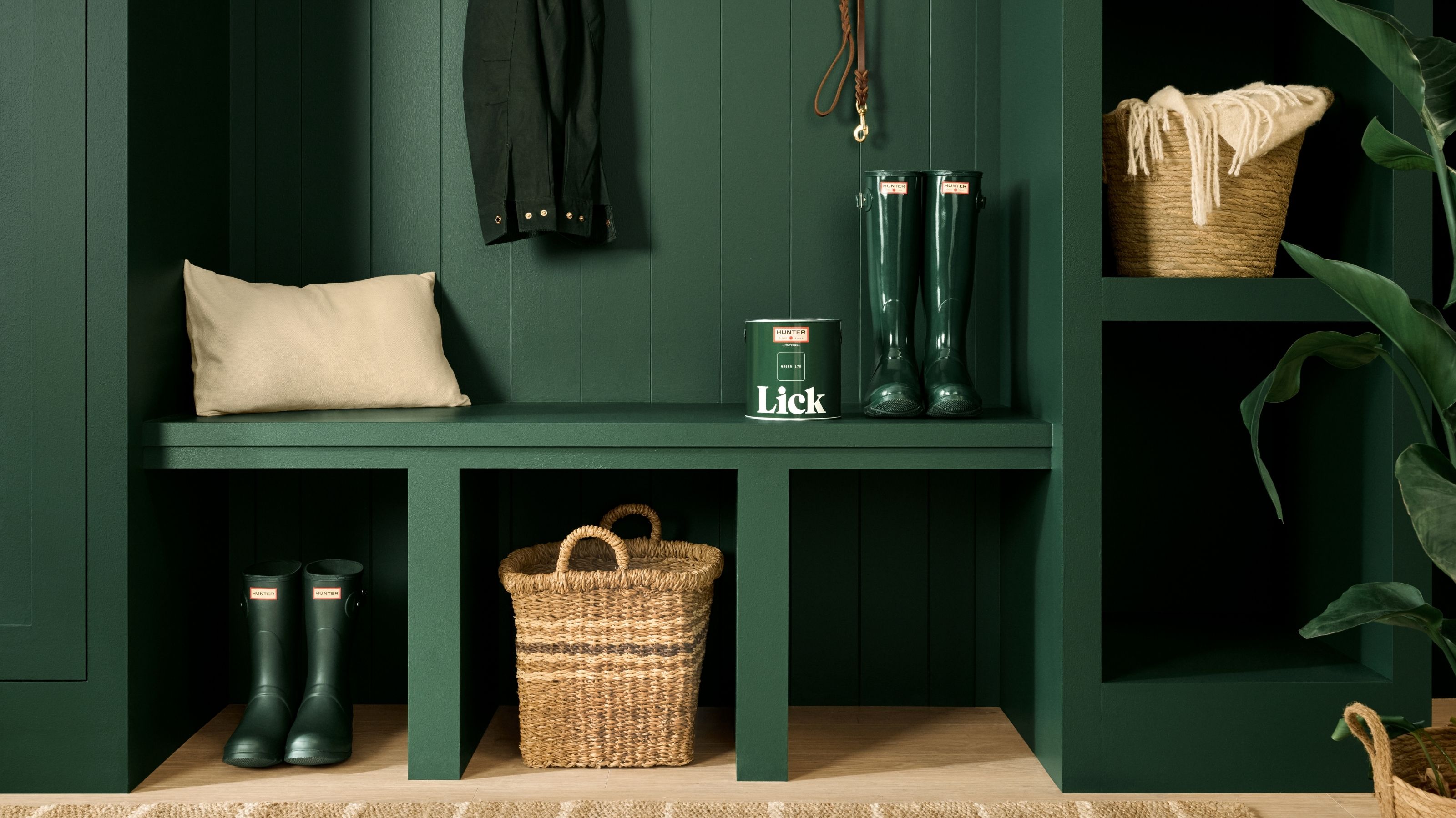 Image of a hunter green built-in mud room unit. There are two pairs of hunter green hunter boots, woven baskets, a can of Lick paint, a beige pillow, and a jacket hanging.