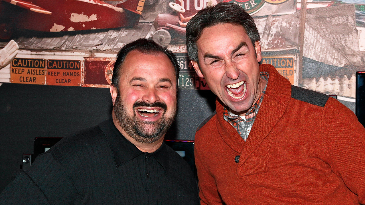 Getty image of Frank Fritz wearing black and Mike Wolfe wearing orange in an early American Pickers promo photo. Both are doing goofy faces.