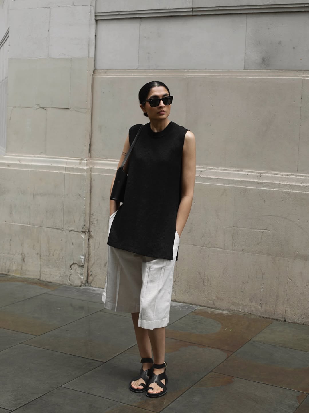 @by.yatri wears a black sleeve-less knit, Bermuda shorts and sandals