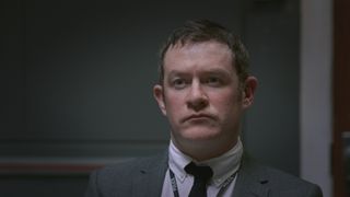 Philip Hill-Pearson as Detective Bell looking serious and stern in a grey suit and grey interrogation room.