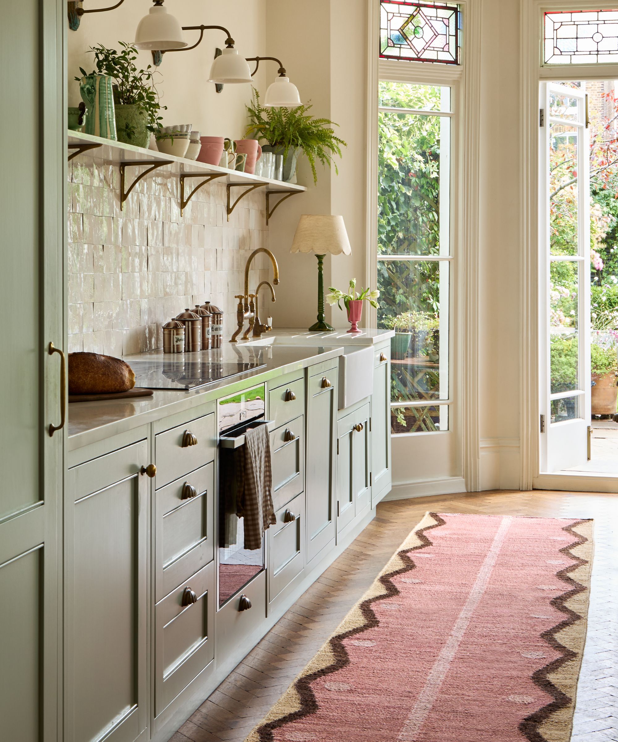 Kitchen with sage green painted cabinetry, cream backsplash, open shelving with potted plants, parquet flooring, a pink and jute runner, and French doors leading out onto the garden