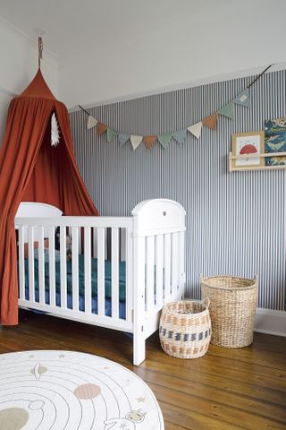 Child's bedroom with blue and white pinstripe wallpaper, white cot bed with terracotta bed canopy, rattan toy storage baskets and bunting