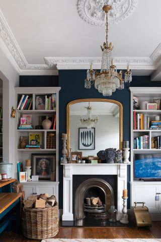 A living room with blue wall paint decor, luxury chandelier light fixture, brass-framed large mirror above fireplace and bookshelves either side of fireplace