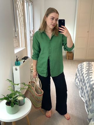 Woman in mirror wears green shirt, black linen trousers and basket bag