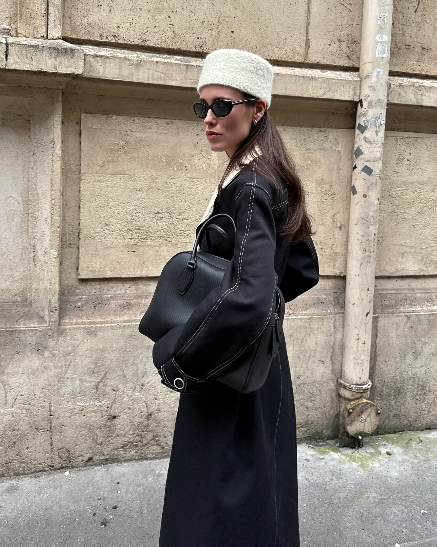 Woman wears white hat, black coat, carries black The Row India bag