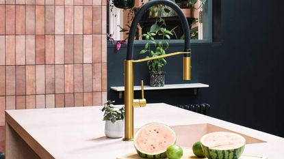A brass-colored tap/faucet in kitchen with pink tile decor and watermelon and lime fruits on kitchen countertop
