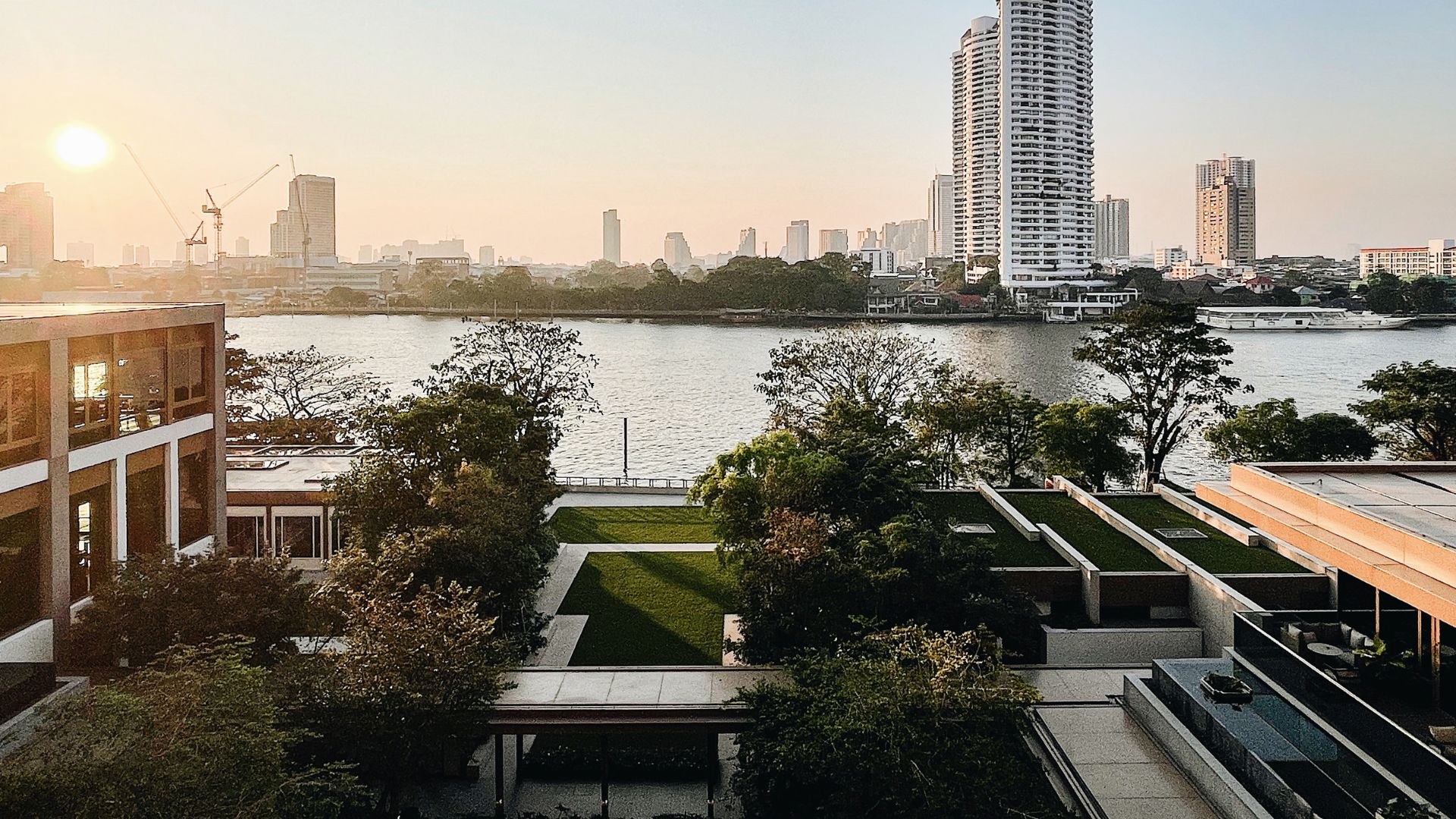 The exterior of the capella bangkok hotel looking out on the river, with skyscrapers and a sunset in the distance
