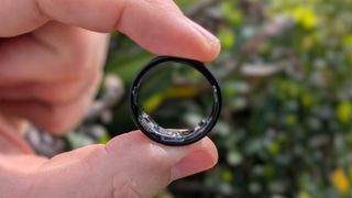 Close-up photo of the black version of Samsung's Galaxy Ring held between thumb and index finger in front of a garden.