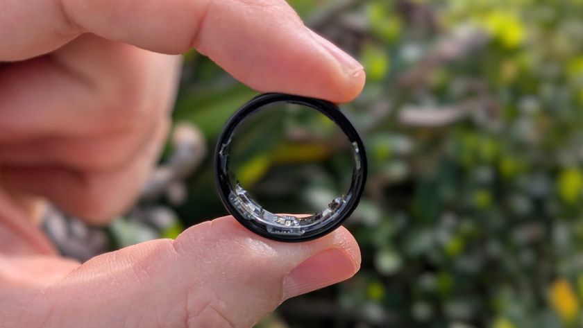 Close-up photo of the black version of Samsung&#039;s Galaxy Ring held between thumb and index finger in front of a garden.