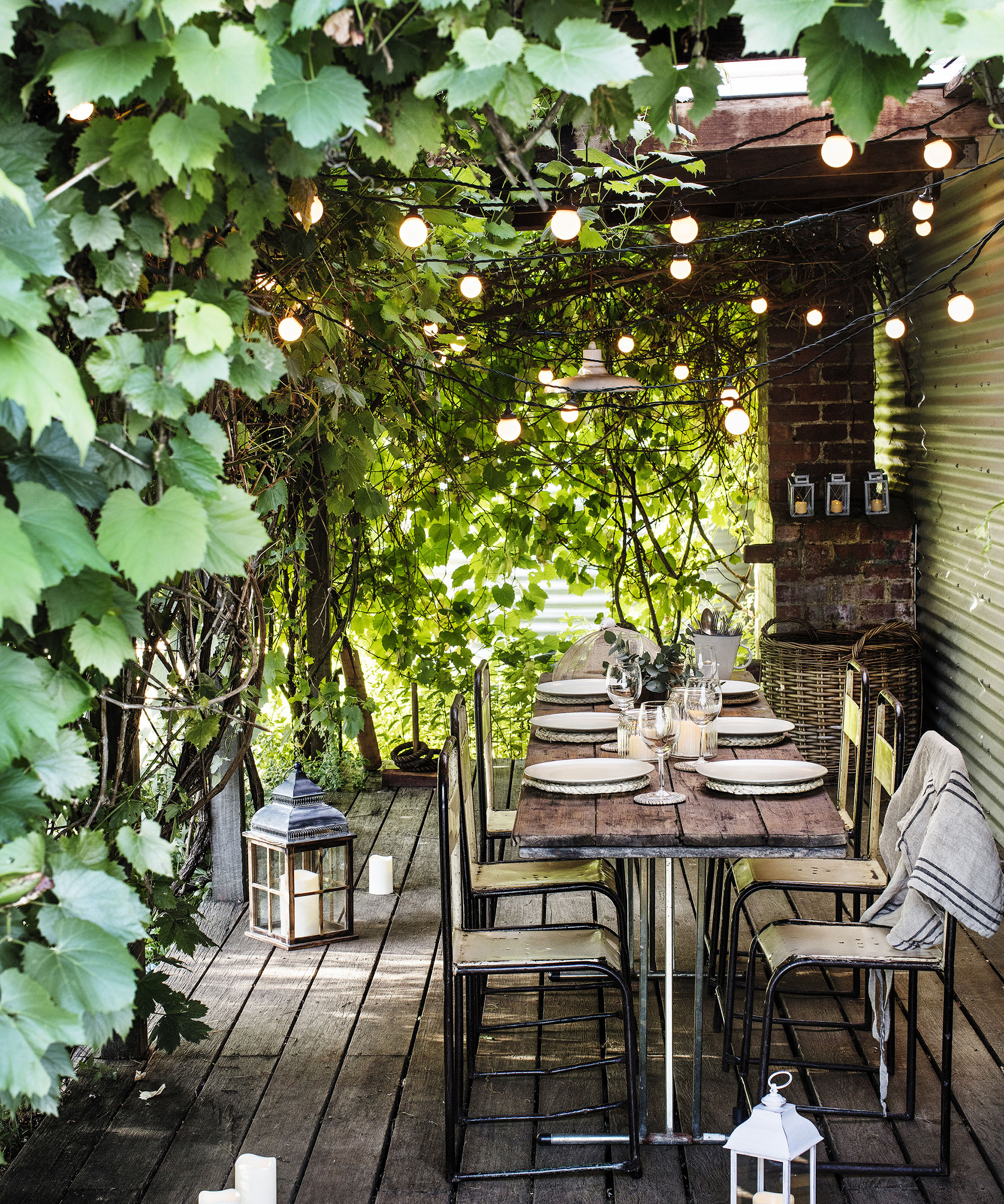 An outdoor table under a shaded archway with climbing plants and festoon lights