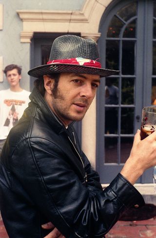 View of Joe Strummer wearing a black and red hat and a black jacket during the day. He is holding a glass of drink and there is a young person in the background wearing a white t-shirt