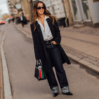 Fashion week attendee wearing sunglasses, button down shirt, black coat, cuffed jeans 