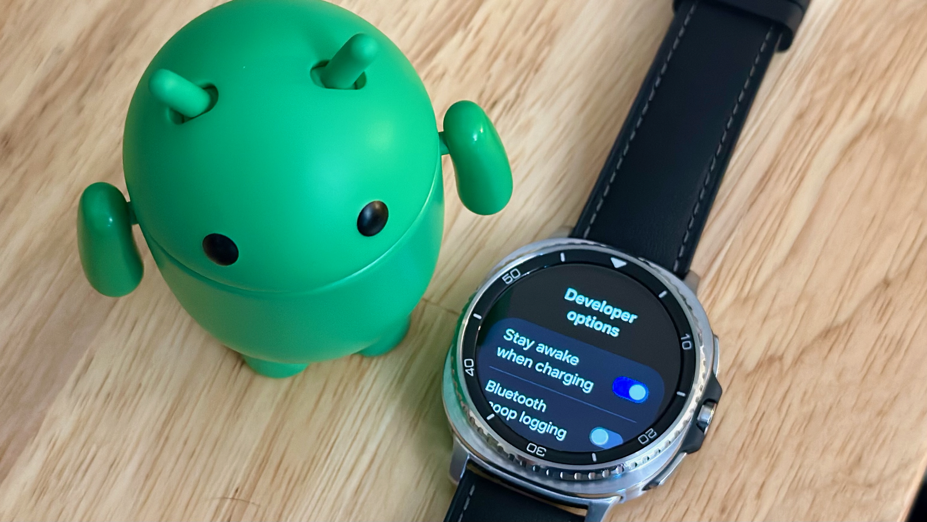 The Samsung Galaxy Watch 8 Classic sitting next to an Android figurine, the display showing "Developer options."