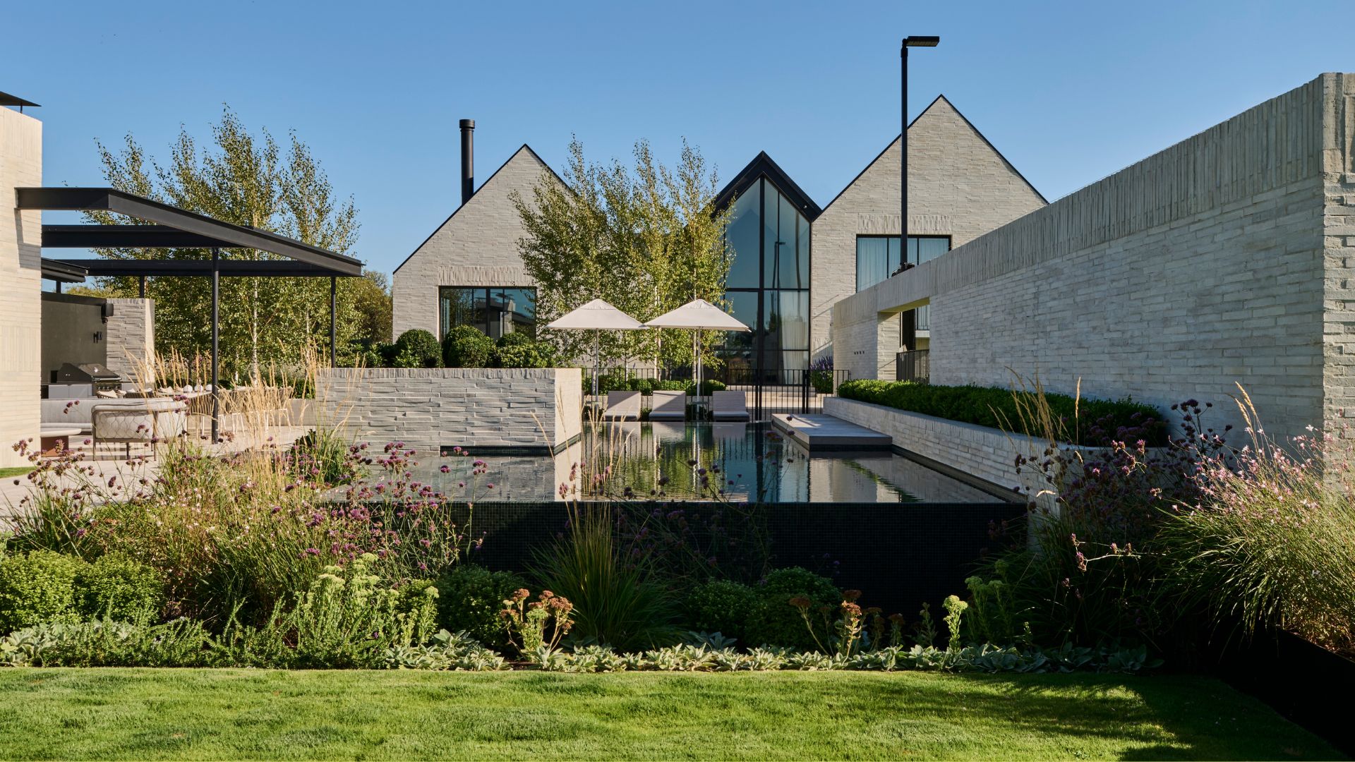 An outdoor garden with a pool, an outdoor living room, a green lawn, tall feathered purple flowers, and ground cover