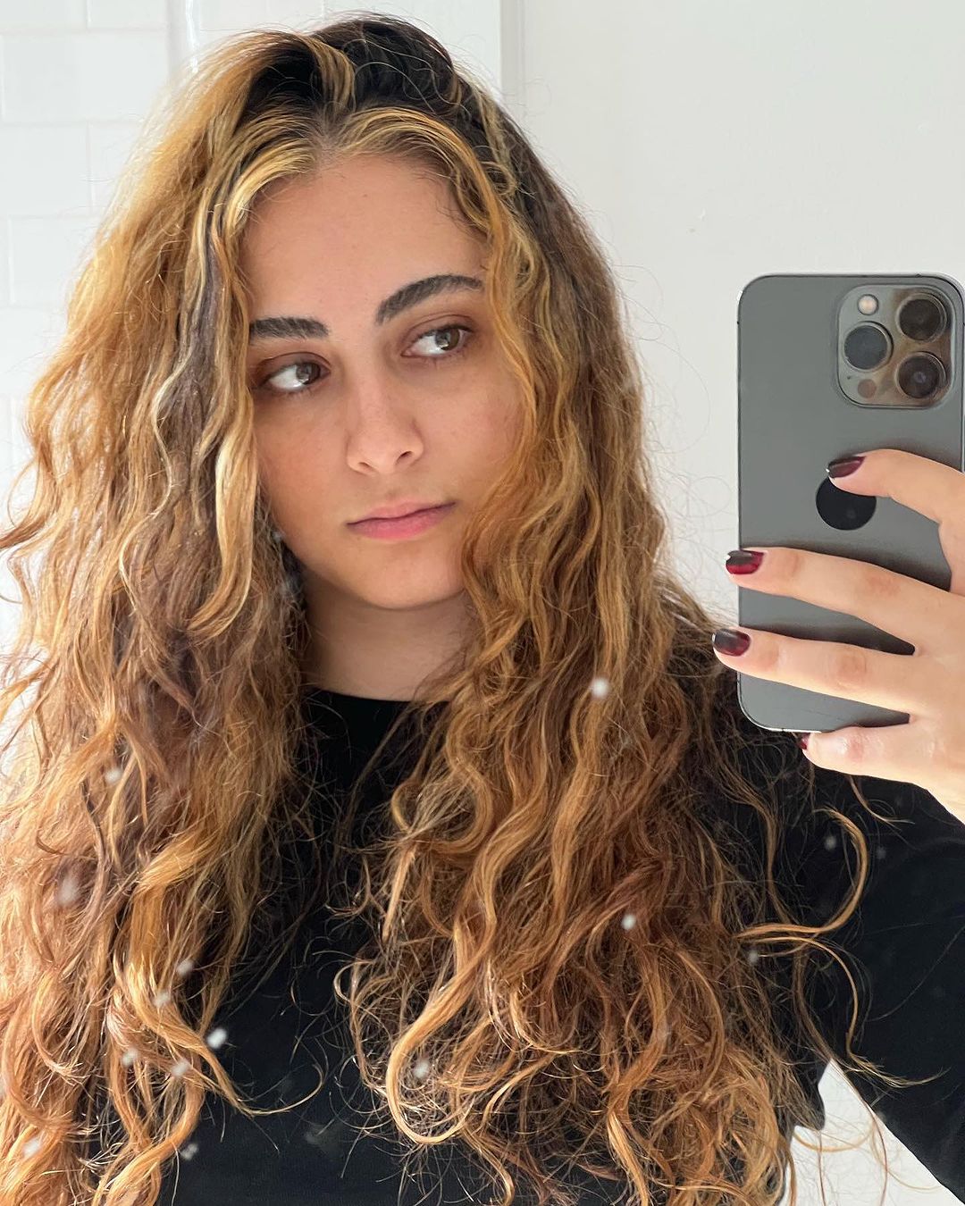 A woman taking a photo with her phone in the mirror with long curly hair and face-framing highlights.