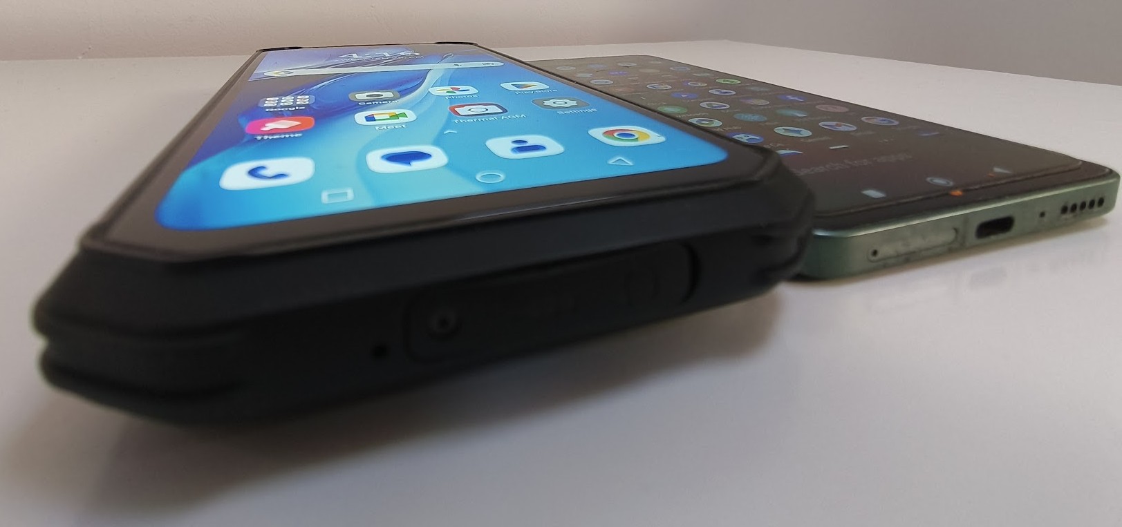 A side-by-side comparison shows a thick, rugged smartphone with a protective black casing next to a much slimmer, green smartphone lying on a white surface.