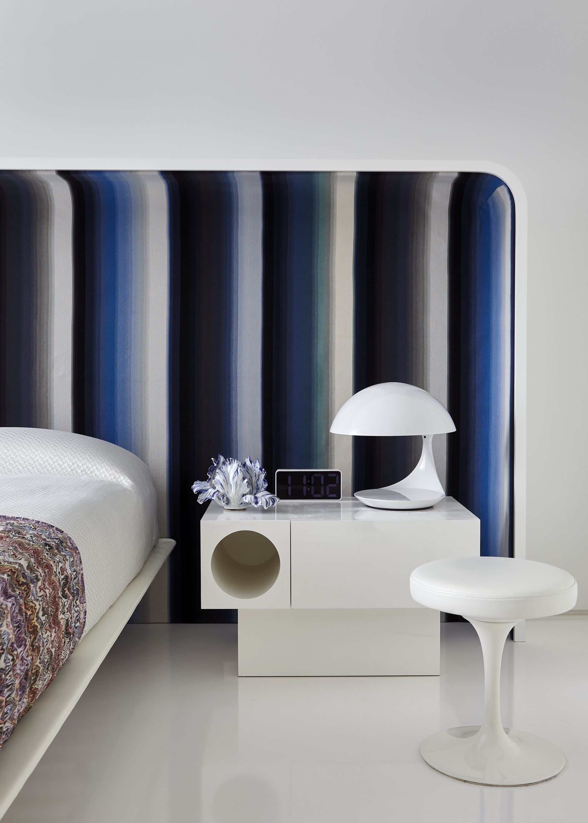 Image of a white bedroom with a minimalistic bed that has an extended headboard. The headboard is a blue and black multi-striped design. There is a white, cube bedside table with a white mushroom lamp on the table.