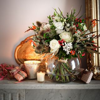 christmas flower arrangement in a glass fishbowl sitting an a grey marble mantle alongside a gold tray lit up by candles