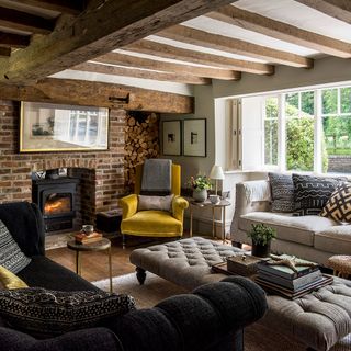 Living room with exposed wooden beams on the ceiling, exposed brick wall around a fireplace and cosy sofas and armchairs around a long cushioned footstool