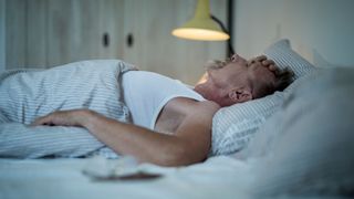 A man in a white vest lays on his back in bed with one hand placed against in forehead in anguish