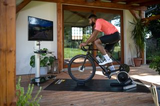 This image shows a rider on his bike, which is mounted on a turbo looking at a screen displaying a Rouvy ride. He is situated in a house with wooden floors and a large window and plants in the background.