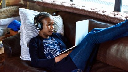 A young woman wears headphones while lying on the sofa with her laptop on her lap.