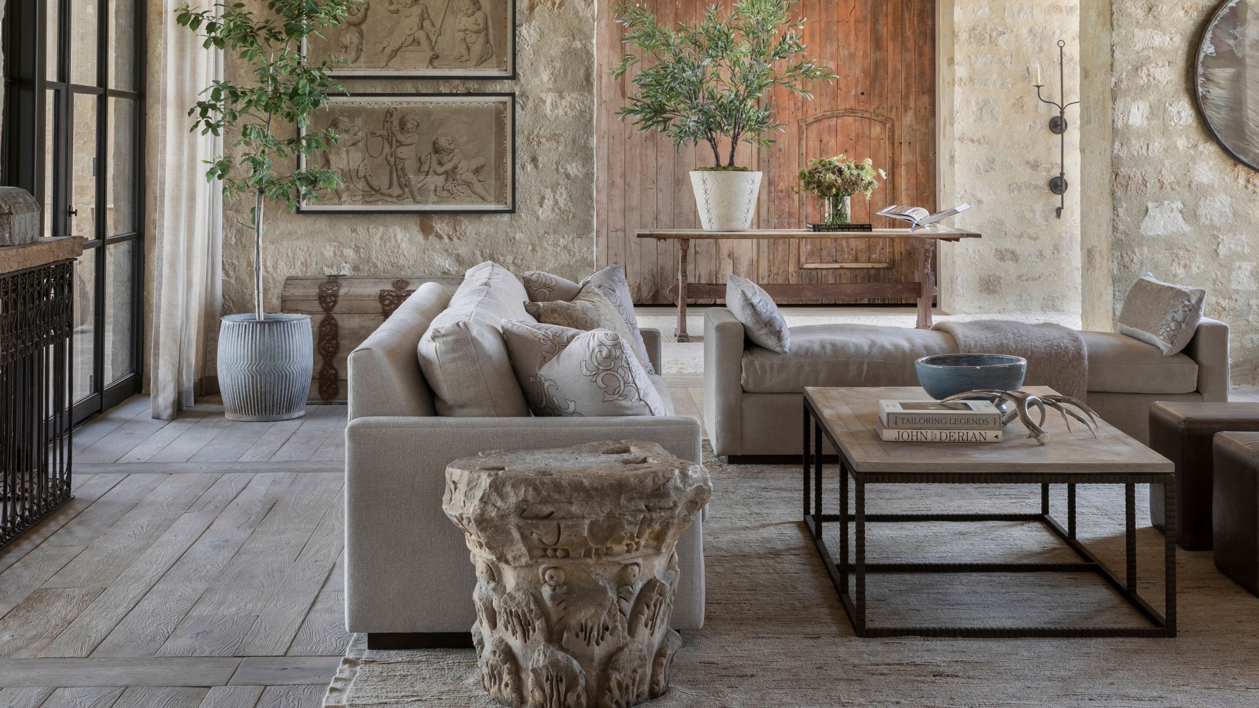 Rustic living room with stone walls, neutral fabric sofas, a carved stone pillar end table, potted indoor trees, and a wooden coffee table with books.