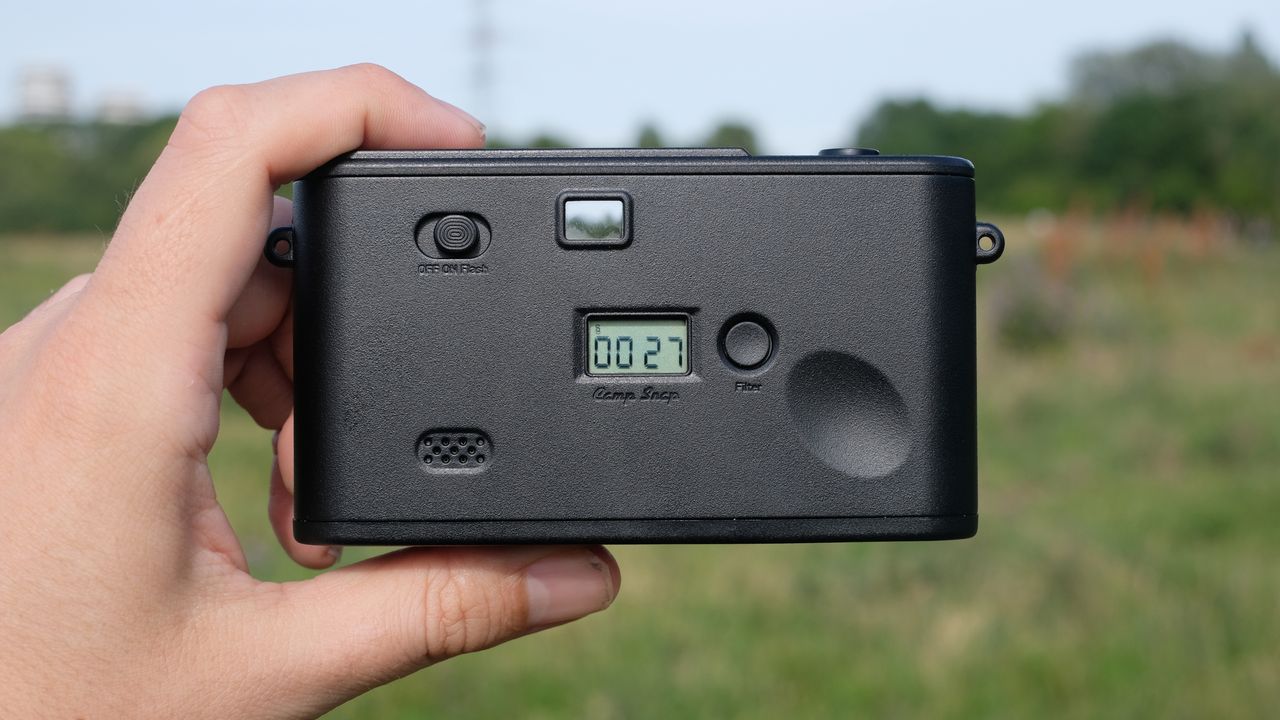 Photograph of man's hand holding Camp Snap 2 digital camera, in yellow colorway, rear view showing shot counter LCD, speaker, filter button and power switch